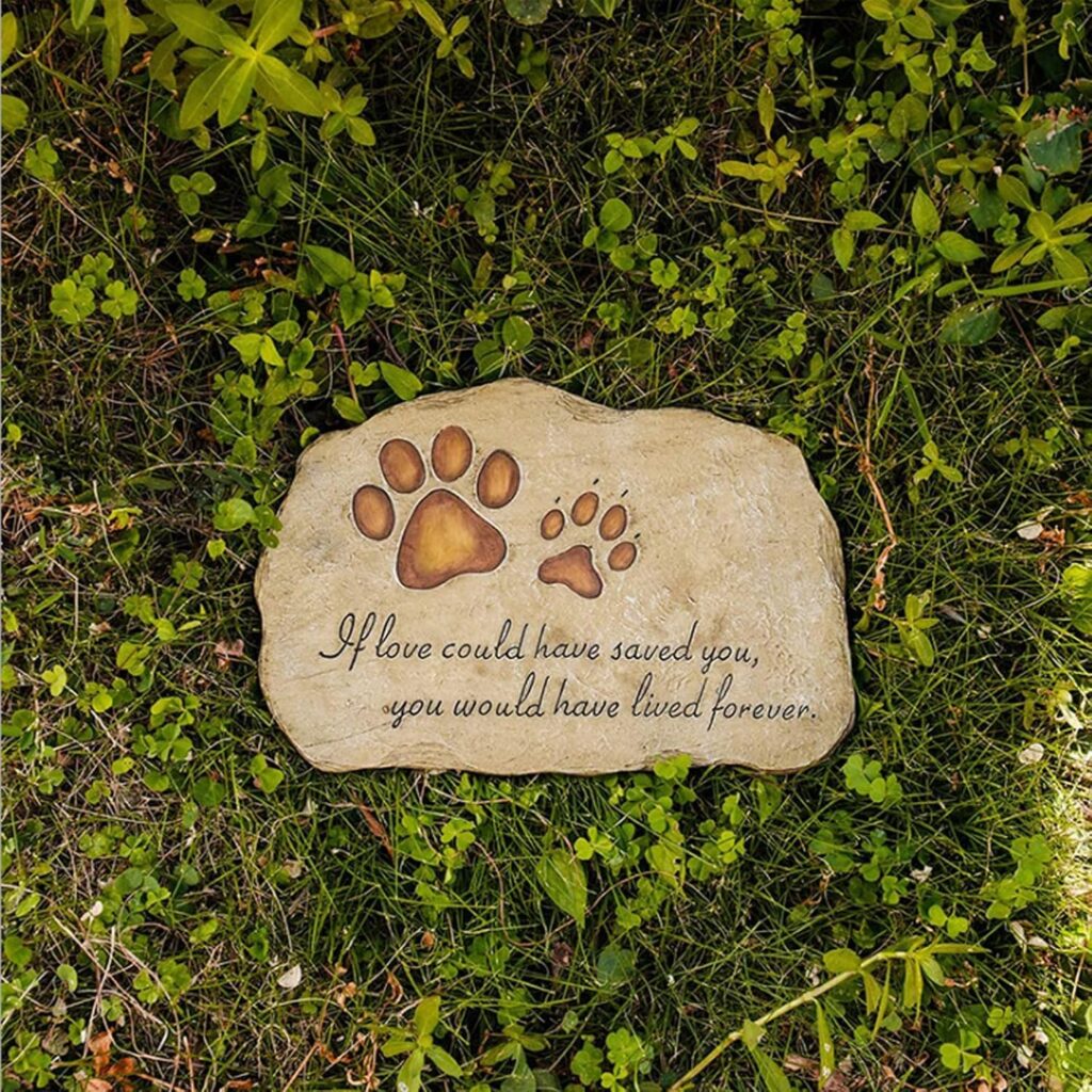 Una piedra conmemorativa situada en el centro de un jardín sobre césped verde y tréboles. La piedra tiene grabadas dos huellas de perro y la frase en inglés: "If love could have saved you, you would have lived forever". La luz natural resalta la textura de la piedra y el verde vibrante de las plantas alrededor.
