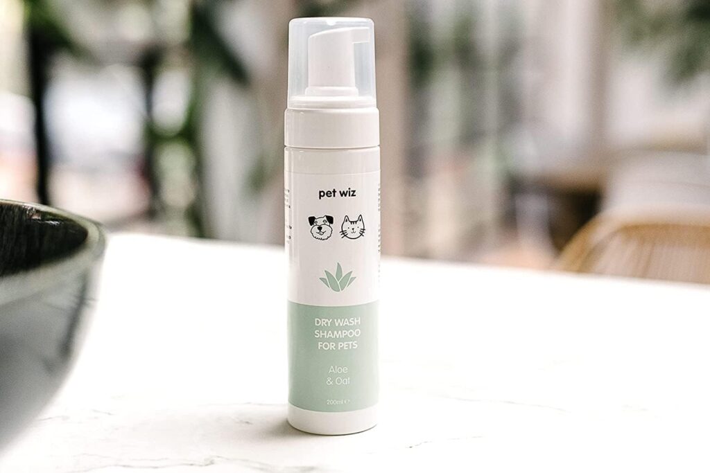 sp1.1 Botella de 200ml de champú en seco "Dry Wash Shampoo" para mascotas de la marca Pet Wiz. El envase es blanco con una franja verde menta en la base, mostrando iconos de un perro, un gato y una planta de aloe vera. La etiqueta indica que contiene Aloe y Avena (Aloe & Oat). El bote tiene un dispensador de espuma con tapa transparente y está situado sobre una encimera de mármol claro.