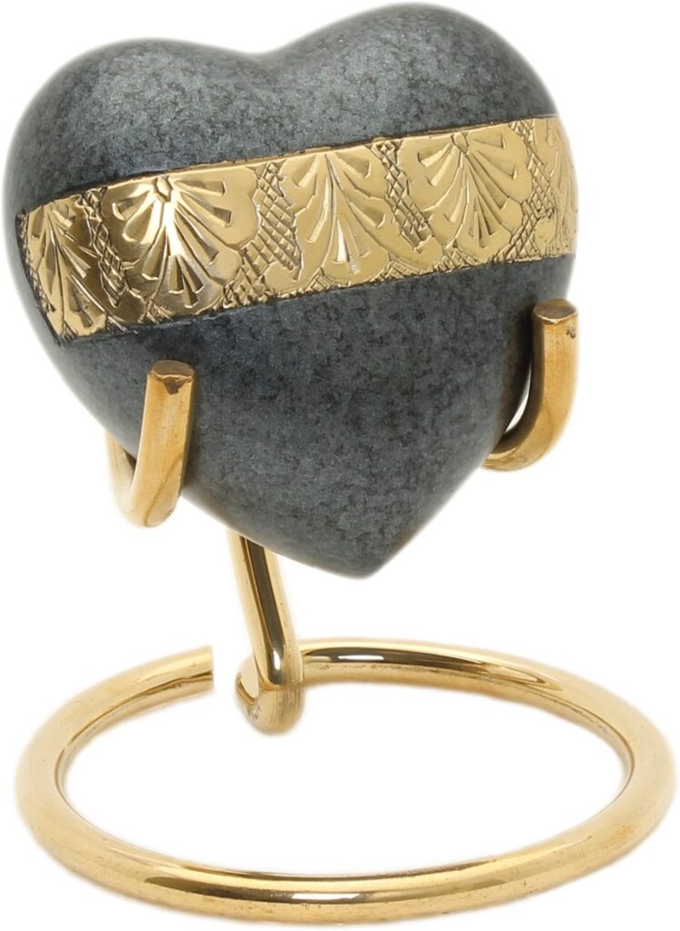 Urna funeraria pequeña en forma de corazón con acabado de piedra gris moteada. Presenta una banda central de metal dorado grabada con motivos ornamentales y se apoya sobre un soporte metálico dorado minimalista.